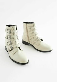 Leather ankle boots in off-white with three adjustable straps, silver buckles, and stud accents. Rounded toe and a low black heel.