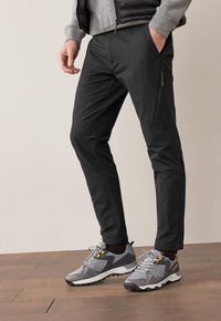 Musta, slim-fit housut joustavasta materiaalista, joissa on vetoketjullinen tasku sivulla ja sileä pinta, yhdistettynä harmaisiin lenkkareihin.
