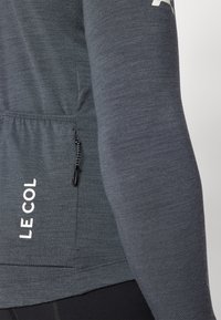 Haut de sport à manches longues gris foncé avec une poche zippée, tissu texturé et logo "LE COL" en blanc sur le côté. Cordons de serrage ajustables.
