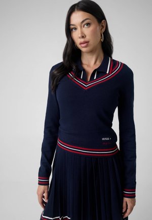Kvinde med langt mørkt hår iført en navyblå sweater og plisseret nederdel med rød- og hvidstribet kant, står mod en ensfarvet baggrund.