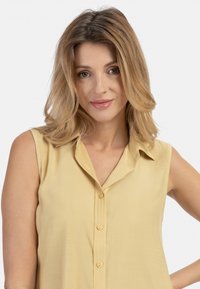 Mouwloze gele blouse met een kraag, voorzien van een knoopsluiting aan de voorkant en een gladde textuur. Lichte stof valt soepel om de schouders.