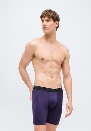 Jeune homme aux cheveux bruns courts portant un boxer Puma violet foncé, debout avec la main gauche derrière le dos, regardant vers sa droite.