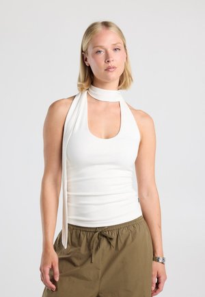 Vrouw met steil blond haar draagt een mouwloze witte haltertop met sjaaldetail en olijfgroene broek met trekkoord, staand tegen een effen achtergrond.