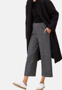 Weit geschnittene, graue karierte Hose kombiniert mit einem schwarzen strukturierten Mantel und weißen Sneakern. Der Mantel hat ein langes, offenes Design.