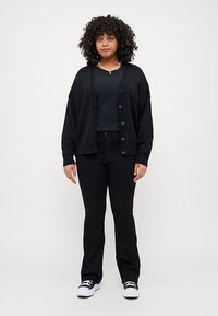 Svart kviltet cardigan med lange ermer, knappefront, brukt over en svart topp, kombinert med svarte vide bukser og svart-hvite sneakers.