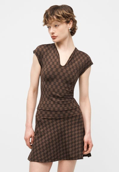 Νέα γυναίκα με κοντά μαλλιά φορώντας καφέ μπλούζα με σχέδια και V-λαιμόκοψη και ταιριαστή φούστα, στέκεται με τα χέρια χαλαρά μπροστά σε λευκό φόντο.