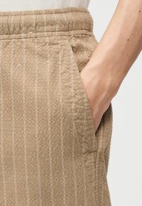 Χέρι τοποθετημένο στην τσέπη μπεζ υφασμάτινου παντελονιού με κάθετες λευκές ρίγες και ελαστική μέση.