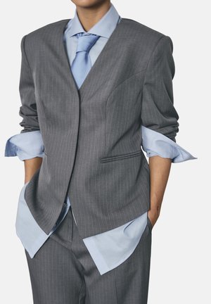 Person iført gråt jakkesæt med lyseblå skjorte og slips, skjortærmerne er rullet op, hænderne i lommerne, billedet er beskåret ved halsen.