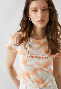 Witte t-shirt met oranje tie-dye patroon, korte mouwen, ronde halslijn en tekst "HET IS NOOIT TE LAAT VOOR FUN" in vetgedrukte oranje letters.