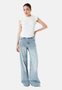 Blusa blanca texturizada con mangas acampanadas combinada con jeans de pierna ancha de color azul claro. El conjunto presenta un ajuste relajado y un mínimo de detalles.