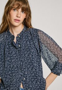 Marinebluse mit weißem Blumenmuster, gerafftem Ausschnitt und transparenten Ärmeln. Verfügt über eine Knopfleiste und leichte Rüschendetails.