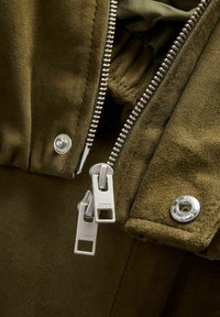 Gros plan sur une veste en daim vert olive avec une fermeture éclair double argentée partiellement ouverte et des boutons-pression métalliques sur le col.