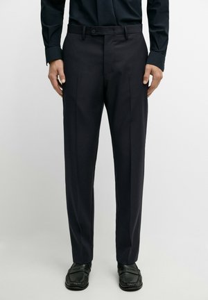 Man draagt zwarte nette broek, zwart net overhemd, zwarte stropdas en zwarte leren loafers, staand tegen een eenvoudige lichte achtergrond.