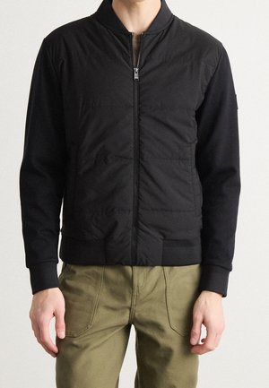 Mężczyzna w czarnej pikowanej kurtce na zamek z prążkowanymi mankietami i kołnierzem, w połączeniu z oliwkowozielonymi spodniami.