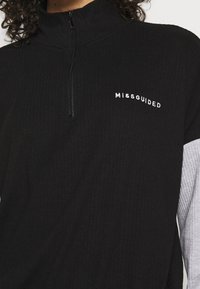 Personne portant un pull côtelé noir avec une fermeture éclair au quart, un logo blanc « MISSGUIDED » sur la poitrine et des manches grises en dessous.