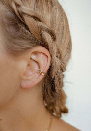 Gros plan sur une oreille avec un manchon en or sur le cartilage orné de petites pierres blanches, cheveux tressés clairs tirés en arrière sur un fond uni.