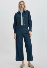 Denimjack met een donkerblauwe wassing, twee borstzakken en gouden knopen, gecombineerd met bijpassende wijde broeken en een lichtgroene trui.