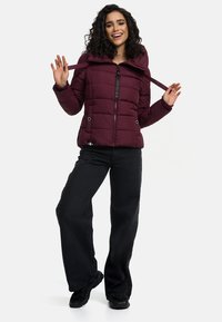 Bordeaux pufferjack met hoge kraag, voorkant rits en verstelbare linten, gecombineerd met zwarte wijde jeans en sneakers.