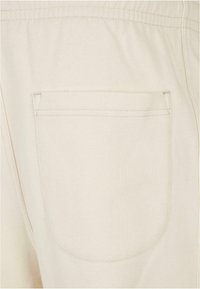 Pantaloni din bumbac bej, având un singur buzunar posterior cu margini cusute. Textura este netedă, cu un model țesut subtil.