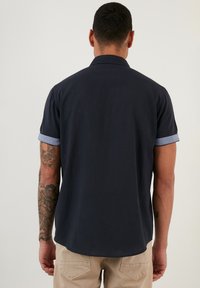 Cămașă scurtă de culoare bleumarin închis, cu guler elegant și manșete răsfrânte de un albastru deschis, confecționată dintr-un material fin, ușor și având un tiv curbat.