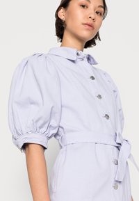 Vestido de ganga lilás claro com frente abotoada, mangas bufantes e cinto amarrado na cintura. Possui botões prateados e bolsos laterais.