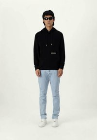 Felpa nera con cappuccio e lacci, tasca frontale e logo bianco. Jeans blu chiaro e sneakers bianche. Il modello indossa occhiali da sole neri.