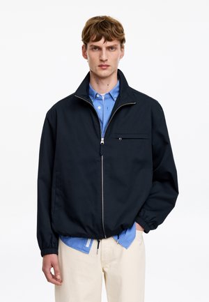 Jeune homme portant une veste noire zippée sur une chemise bleue à boutons et un pantalon beige, debout devant un fond blanc uni.