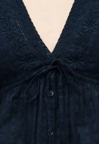 Κοντινό πλάνο φορέματος από υφασμάτινη μπλε σκουριά με υφή, κεντήματα με λουλούδια, δαντελωτό μπούστο σε σχήμα V, κουμπιά μπροστά και δεμένη φιόγκο στο στήθος.