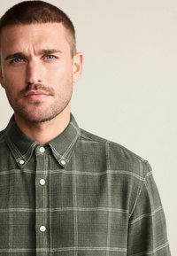 Camisa a cuadros verdes con cuello abotonado, acentos blancos y una tela texturizada. Presenta botones blancos en la tapeta delantera.