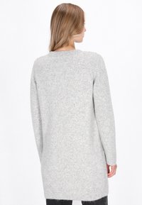 Langer, hellgrauer Pullover mit einem strukturierten Strickmuster. Er hat einen runden Halsausschnitt und gerippte Bündchen. Die Rückseite ist schlicht und reicht bis zur Mitte des Oberschenkels.