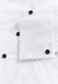 Balts krekls ar melniem pogām, kas izceļas ar tīru aproci un gludu tekstūru. Audums izskatās viegls un ir sakārtots.