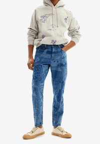 Sudadera con capucha gris con gráficos morados, combinada con jeans de mezclilla azul con estampados impresos y zapatillas beige con suelas texturizadas.