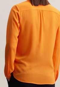 Long-sleeve shirt in bright orange, made of smooth fabric. Features a rounded hem, buttoned cuffs, and a subtle pleat at the back.