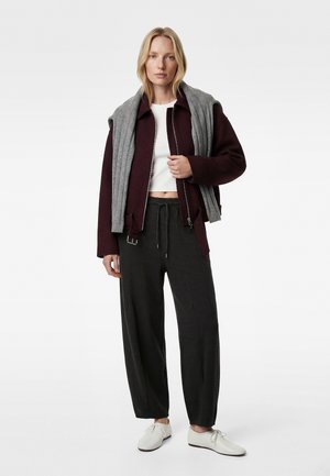 Donna in piedi che indossa una giacca bordeaux, un top corto bianco, pantaloni neri con coulisse, scarpe bianche e un maglione grigio appoggiato sulle spalle.
