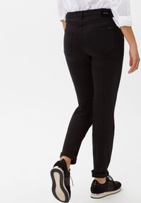 Pantalones de mezclilla negros con un corte ajustado, que cuentan con bolsillos traseros y una etiqueta de logo. Combinados con zapatillas negras, enrolladas en el tobillo.