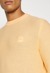 Närbild av en person som bär en persikofärgad texturerad stickad tröja med en liten fyrkantig "BOSS" logotyp patch på bröstet.