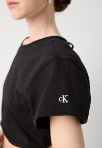 Fekete, rövid ujjú, puha anyagból készült, cropped póló, amelyen a bal ujján fehér hímezett "cK" logó található.
