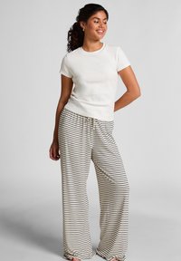 Vrouw met krullend haar in een witte top met korte mouwen en een crème wijdvallende broek met zwarte horizontale strepen, staande tegen een effen achtergrond.