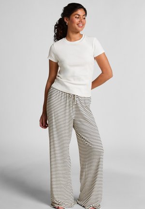 Femme aux cheveux bouclés portant un haut blanc à manches courtes et un pantalon large crème avec des rayures horizontales noires, se tenant devant un fond uni.