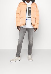 Puffer jacket in soft peach, paired with a white graphic tee, gray jeans, and beige sneakers with black accents. Simple design, casual fit.