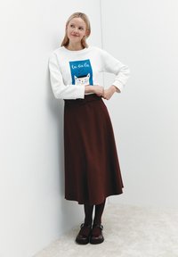 Witte sweatshirt met een kattenafbeelding en blauwe en rode details, gecombineerd met een bruine rok tot de knie en zwarte schoenen.