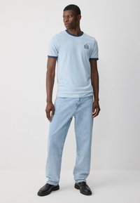 T-shirt en coton bleu clair avec col côtelé bleu marine, arborant un petit logo brodé sur la poitrine gauche, associé à un jean bleu clair et des chaussures noires.