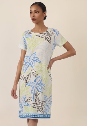 Frau trägt knielanges weißes Kleid mit blau-grünen und braunen Blattmustern, steht mit einer Hand in der Hüfte vor einem beigen Hintergrund.