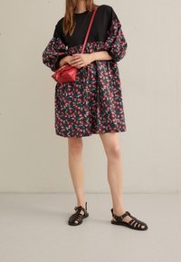 Czarna sukienka z bufiastymi rękawami i kwiatowym wzorem w czerwieni i zieleni; zestawiona z czarnymi sandałami na paski i małą czerwoną torebką na ramię.