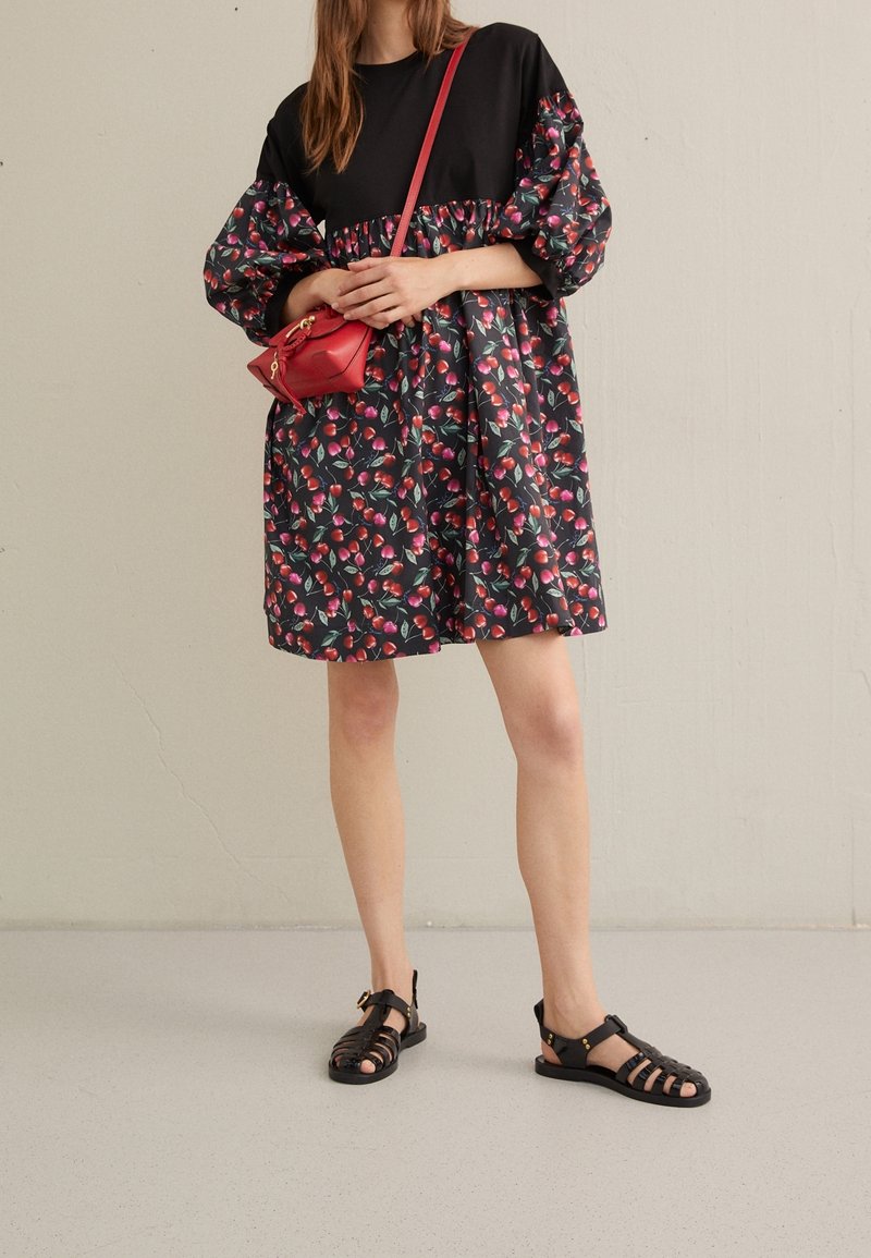 Czarna sukienka z bufiastymi rękawami i kwiatowym wzorem w czerwieni i zieleni; zestawiona z czarnymi sandałami na paski i małą czerwoną torebką na ramię.