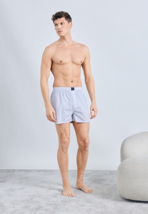Jonge man staat blootvoets op een grijs tapijt naast een witte, textuurrijke fauteuil, dragen lichtblauwe en witte gestreepte boxershorts.