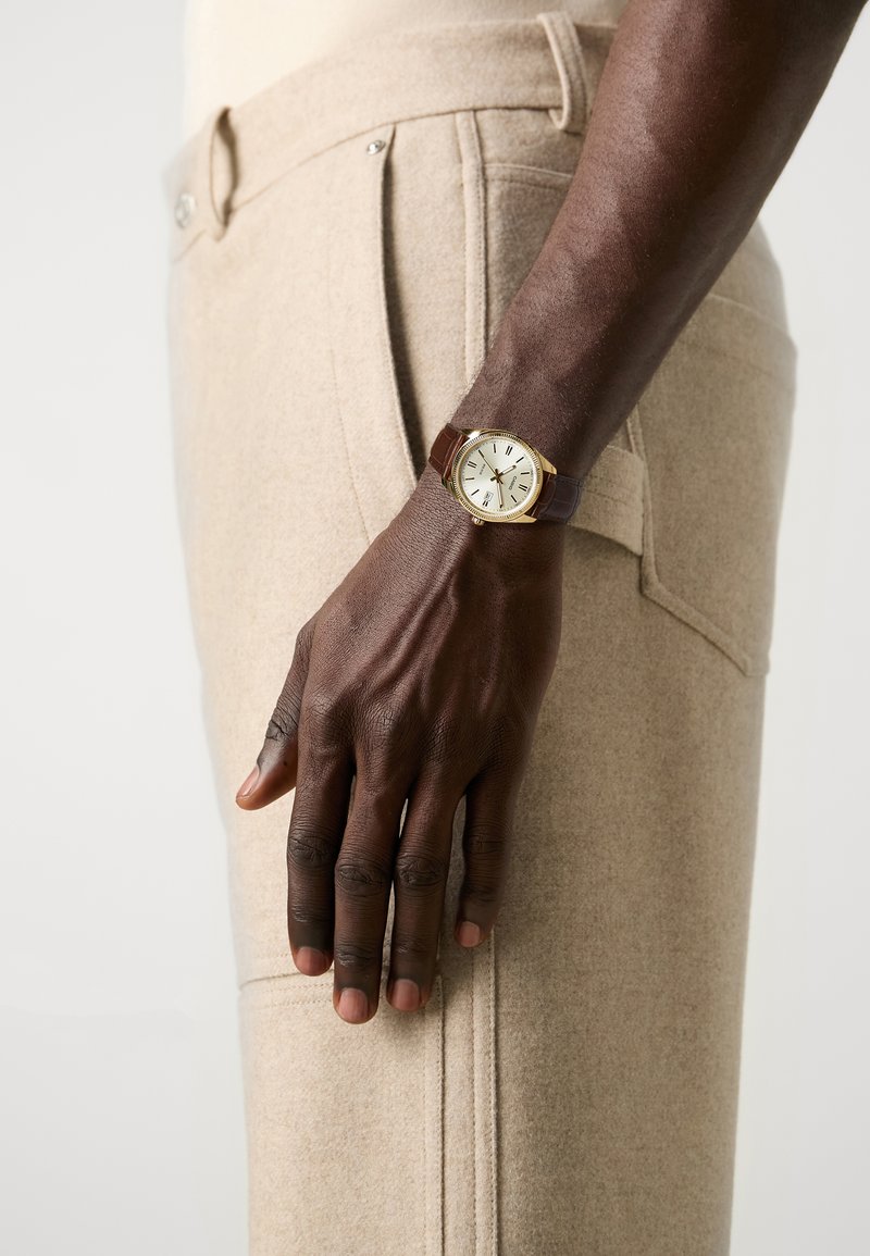 Brown leather strap watch with a white dial and gold-tone casing, worn on a hand beside light beige wool trousers with side pocket detail.