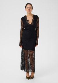 Mustmustrits musta pikkade, läbipaistvate varrukatega ja sügava V-kaelusega. Omab keerukaid lillemustreid ja kokkuhoidvat siluetti, koos pikema mustriga overlay'ga.