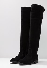 Bottes noires montantes au-dessus du genou en daim souple. Elles présentent un bout arrondi, un talon plat et un design sans coutures avec une fermeture éclair latérale discrète.