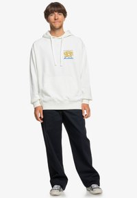 Sudadera blanca de algodón con cordones, bolsillo frontal y un diseño gráfico de olas en dorado y azul. Combinada con pantalones oscuros y zapatos grises.